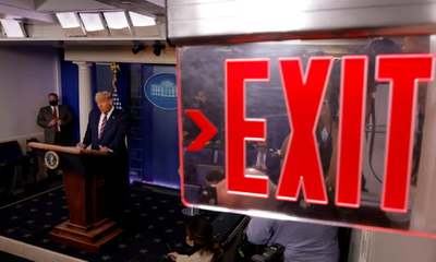 Donald Trump at the White House briefing room podium, seen past a glowing red exit sign.
