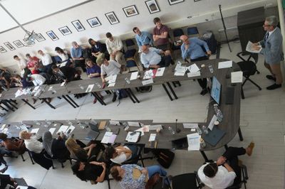 Overhead view of a conference room, with people around a U-shaped table and a speaker standing at one end.