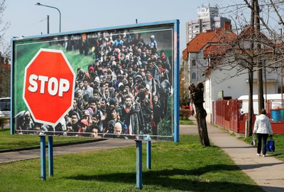 Roadside government billboard with a red STOP sign over a crowd of migrants, beside a quiet residential street.