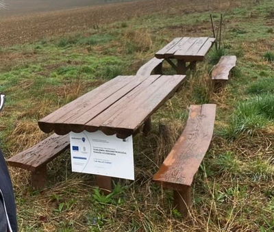 Two rough wooden picnic tables and benches in a muddy field, with an EU project sign fixed to one.