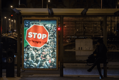 Government poster at a bus stop at night, showing a red STOP sign over a crowd of migrants.